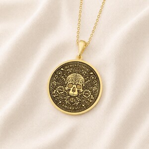 Gold tone Memento Mori necklace on cream fabric with an engraved skull, roses, hourglasses, scrolling leaves, and Amor Fati inscription.