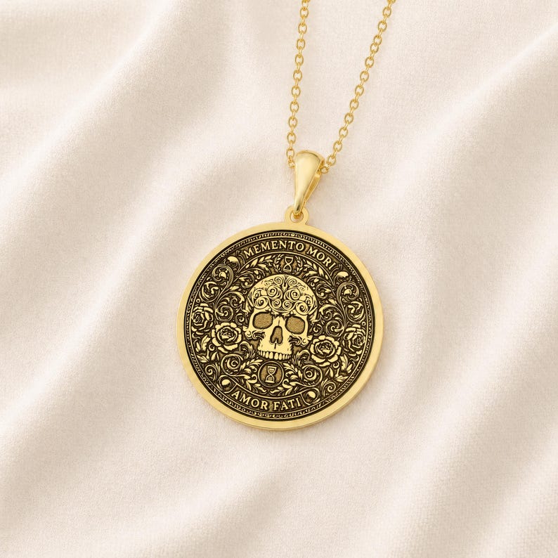 Gold tone Memento Mori necklace on cream fabric with an engraved skull, roses, hourglasses, scrolling leaves, and Amor Fati inscription.