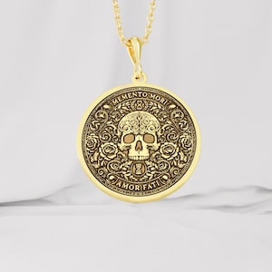 Gold tone Memento Mori necklace on white fabric with an engraved skull, roses, hourglasses, scrolling leaves, and Amor Fati inscription.