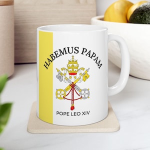 May include: White ceramic mug with a yellow stripe and the words "HABEMUS PAPAM" and "POPE LEO XIV" printed on it. The mug features a detailed emblem with a crown, keys, and a red cord. The mug is sitting on a beige coaster.
