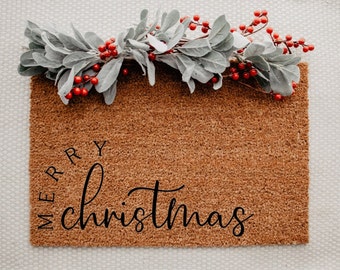 May include: A rectangular brown coir doormat with the words "MERRY christmas" in black script. The mat is decorated with a garland of gray leaves and red berries, perfect for the holiday season.