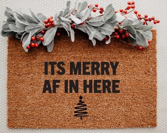May include: A rectangular brown coir doormat with the text "ITS MERRY AF IN HERE" in black. A stylized Christmas tree is below the text. The mat is decorated with a garland of gray leaves and red berries.