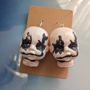 May include: A pair of earrings featuring painted doll heads with black and white markings. The doll heads are pink and have a creepy, doll-like appearance.