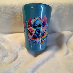 May include: A blue tumbler with a glittery finish, featuring a cartoon image of Stitch surrounded by flowers and a rainbow. The tumbler has a slightly tapered shape. The design is vibrant with shades of blue, pink, and yellow.