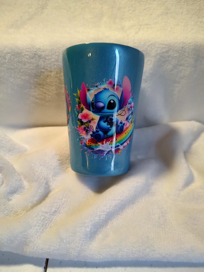 May include: A blue tumbler with a glittery finish, featuring a cartoon image of Stitch surrounded by flowers and a rainbow. The tumbler has a slightly tapered shape. The design is vibrant with shades of blue, pink, and yellow.