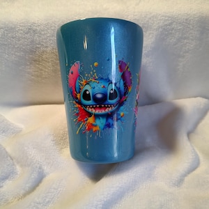 May include: A blue tumbler with a colourful graphic of the character Stitch from Lilo & Stitch. The design includes vibrant splashes of red, yellow, blue, and pink. The tumbler has a glossy finish and is set against a white, textured background.
