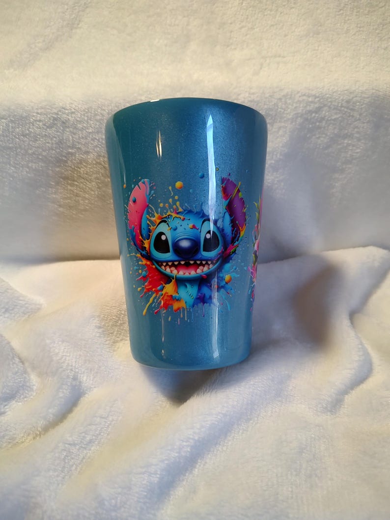 May include: A blue tumbler with a colourful graphic of the character Stitch from Lilo & Stitch. The design includes vibrant splashes of red, yellow, blue, and pink. The tumbler has a glossy finish and is set against a white, textured background.