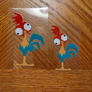 May include: Two cartoon rooster stickers. Each sticker features a rooster with a red comb, large white eyes, a yellow beak and legs, and blue feathers. The stickers are on a clear backing.