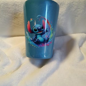 May include: A blue, glitter-finished tumbler, decorated with a cartoon image of Stitch within a vibrant water splash design. The cylindrical tumbler is likely made from a robust material, ideal for cold drinks.