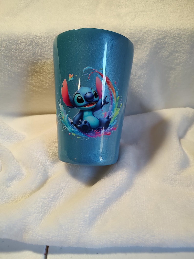 May include: A blue, glitter-finished tumbler, decorated with a cartoon image of Stitch within a vibrant water splash design. The cylindrical tumbler is likely made from a robust material, ideal for cold drinks.