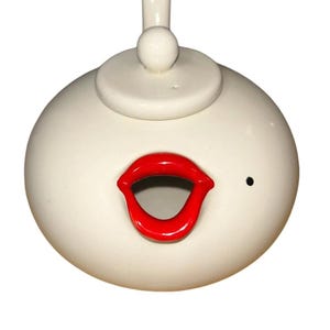 May include: A cream-coloured ceramic teapot with a red lip-shaped spout. The teapot has a rounded body, a small black dot, a lid with a knob, and a curved handle. The teapot is designed to hold hot beverages.