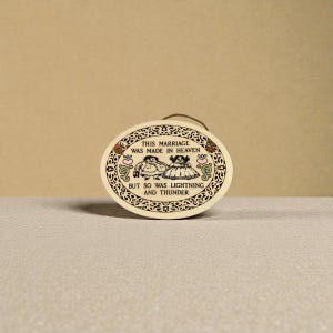 May include: Oval ceramic plaque with a brown handle. The plaque features a black and white illustration of a couple and the text "THIS MARRIAGE WAS MADE IN HEAVEN BUT SO WAS LIGHTNING AND THUNDER". Decorative border with hearts.