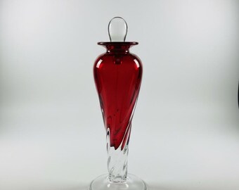 May include: A tall, decorative glass bottle with a red body and a clear, twisted stem. The bottle has a clear, rounded stopper. The base is a clear, circular disc. The bottle is approximately 12 inches tall.