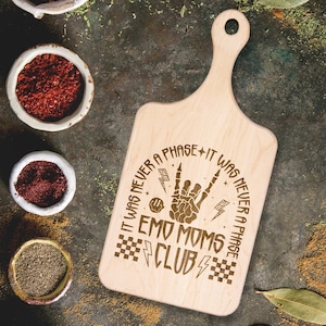 May include: Wooden cutting board with a black and white graphic of a skeleton hand giving the rock and roll sign. The text "It was never a phase + It was never a phase Emo Moms Club" is engraved on the board.