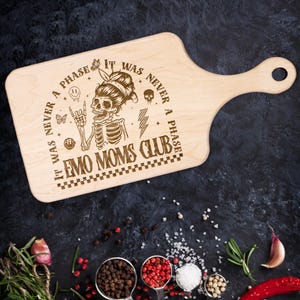 May include: A wooden cutting board with a handle, featuring a laser-engraved design of a skeleton with a punk rock aesthetic. The text reads "It was never a phase. It was never a phase. Emo Moms Club."