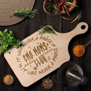 May include: A wooden cutting board with a handle, featuring the text "It was never a phase Emo Moms Club It was never a phase". The text is in a black font and is surrounded by various symbols, including a skull, a butterfly, and a lightning bolt.