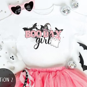 May include: Pink and white girls' Halloween tutu outfit.  The white shirt features a cute graphic that says 'BOO.DAY girl' with pink lettering, bats, and ghosts.  The outfit includes a pink tulle tutu and a pair of pink heart-shaped sunglasses.