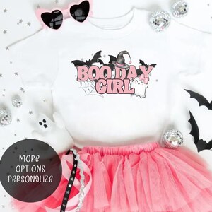 May include: A white t-shirt with a pink graphic that says "Boo Day Girl" with a witch hat, bats, and a ghost. The shirt is paired with a pink tulle skirt and pink sunglasses.