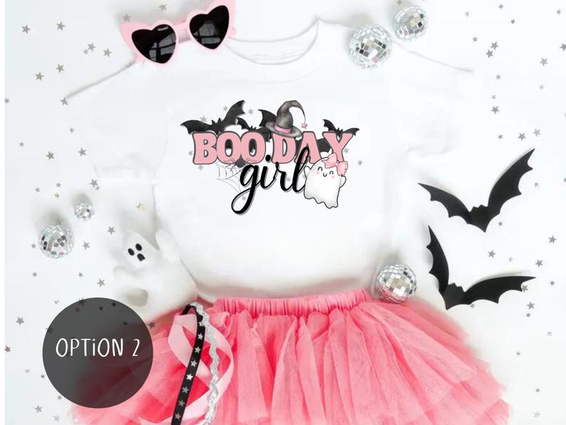 May include: Pink and white girls' Halloween tutu outfit.  The white shirt features a cute graphic that says 'BOO.DAY girl' with pink lettering, bats, and ghosts.  The outfit includes a pink tulle tutu and a pair of pink heart-shaped sunglasses.