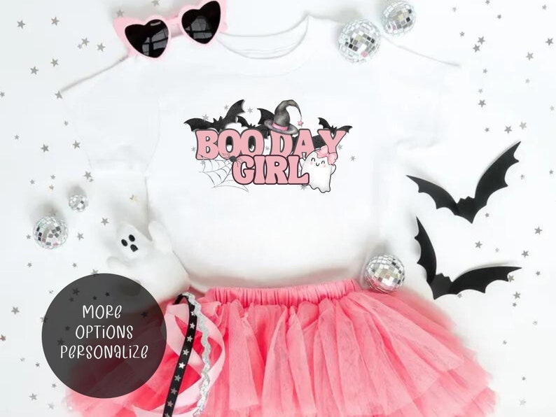 May include: A white t-shirt with a pink graphic that says "Boo Day Girl" with a witch hat, bats, and a ghost. The shirt is paired with a pink tulle skirt and pink sunglasses.
