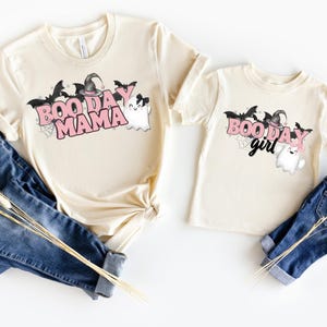 May include: Two cream colored t-shirts with a Halloween theme. The larger shirt says "Boo Day Mama" in pink letters with a ghost and bats. The smaller shirt says "Boo Day Girl" in pink letters with a ghost and bats.