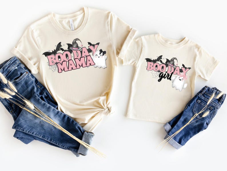 May include: Two cream colored t-shirts with a Halloween theme. The larger shirt says "Boo Day Mama" in pink letters with a ghost and bats. The smaller shirt says "Boo Day Girl" in pink letters with a ghost and bats.