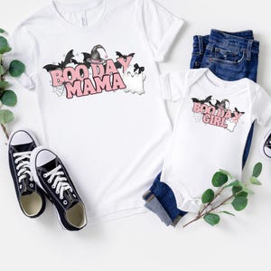 May include: Two white t-shirts with a Halloween theme. The larger shirt says "Boo Day Mama" with a witch hat and bats. The smaller shirt says "Boo Day Girl" with a witch hat and bats.