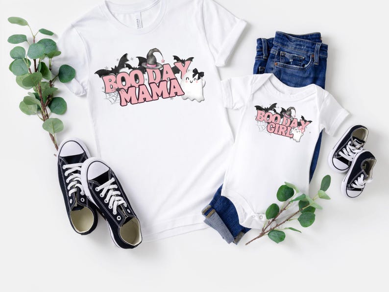 May include: Two white t-shirts with a Halloween theme. The larger shirt says "Boo Day Mama" with a witch hat and bats. The smaller shirt says "Boo Day Girl" with a witch hat and bats.