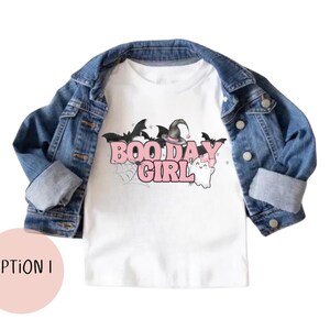 May include: White t-shirt with a pink "Boo Day Girl" graphic featuring a witch's hat, bats, and a ghost. The shirt is layered over a denim jacket.
