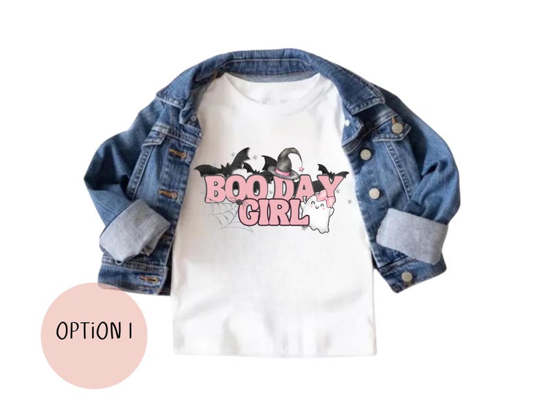 May include: White t-shirt with a pink "Boo Day Girl" graphic featuring a witch's hat, bats, and a ghost. The shirt is layered over a denim jacket.
