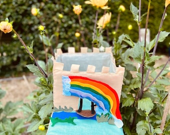 May include: A felt castle playset with a rainbow, clouds, and a small boat. The castle is beige with white trim and is surrounded by green felt grass.
