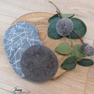 Könnte beinhalten: Wiederverwendbare, runde Abschminkpads in Blau- und Grautönen, einige mit geometrischen Mustern. Die Pads sind auf einer Holzscheibe angeordnet, mit dekorativem Grün.
