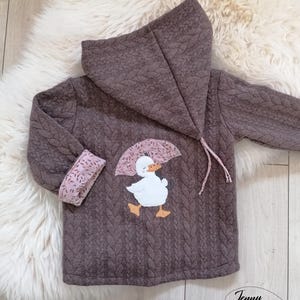 Könnte beinhalten: Braune Kapuzenjacke mit Steppmuster und Entenapplikation mit Regenschirm. Die Manschetten haben ein rosa Blumenmuster. Die Jacke hat eine Kapuze mit Kordelzug und ist für ein Kind konzipiert.