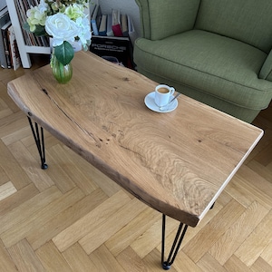 Op de afbeelding: Een rechthoekige salontafel van lichtkleurig hout met zwarte haarspeldpoten. Op de tafel staat een kleine witte kop en schotel met koffie. De tafel is ongeveer 120 cm lang. Er staat ook een vaas met bloemen.