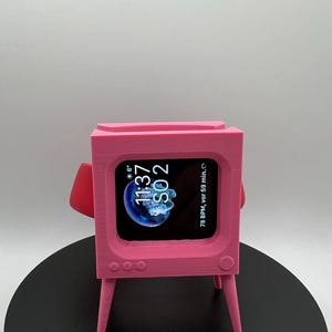 Könnte beinhalten: Ein rosa 3D-gedruckter Ständer in Form eines Retro-Fernsehers mit einem schwarzen Bildschirm, der die Zeit 11:37 und das Datum 80 2 anzeigt. Der Ständer hat vier kleine Kreise auf der Vorderseite und drei Beine. Der Ständer steht auf einer schwarzen Oberfläche.