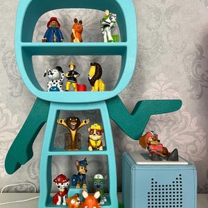 May include: A teal blue cat-shaped bookshelf with multiple shelves filled with various small toys, including a lion, a dog, a fox, and a fireman. A teal blue speaker with a black dot pattern is on the right side of the image.