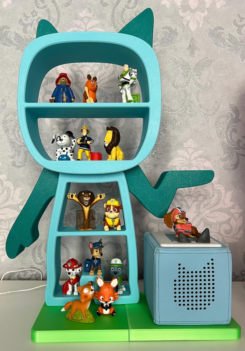 May include: A teal blue cat-shaped bookshelf with multiple shelves filled with various small toys, including a lion, a dog, a fox, and a fireman. A teal blue speaker with a black dot pattern is on the right side of the image.