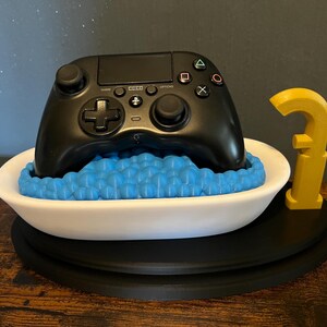 Könnte beinhalten: Ein schwarzer Videospielcontroller, der in einer weißen Badewanne mit blauen Badeschaum liegt. Ein gelber Wasserhahn ist an der Seite der Wanne befestigt. Die Badewanne steht auf einem schwarzen runden Sockel.