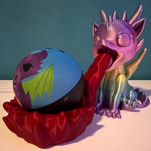 Könnte beinhalten: Eine 3D-gedruckte Drachenfigur mit einem regenbogenfarbenen, irisierenden Finish, die einen blauen und violetten Lautsprecher im Maul hält. Die Zunge des Drachen ist rot und der Lautsprecher hat ein Drachenmotiv.