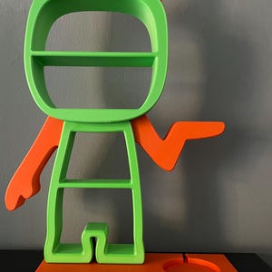 May include: A green and orange shelf unit shaped like a cartoon character with a shelf on the head, two shelves on the body, and a small shelf on the base. The character has two orange ears and one orange arm outstretched.