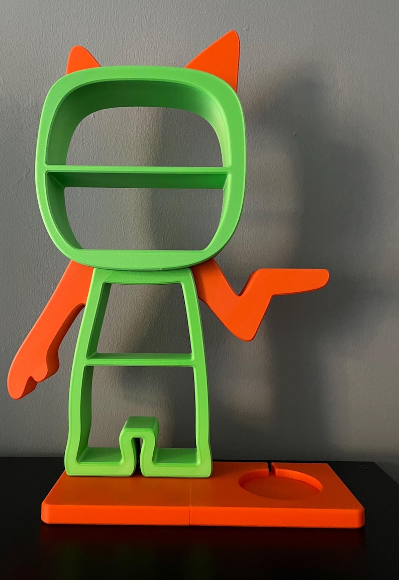 May include: A green and orange shelf unit shaped like a cartoon character with a shelf on the head, two shelves on the body, and a small shelf on the base. The character has two orange ears and one orange arm outstretched.