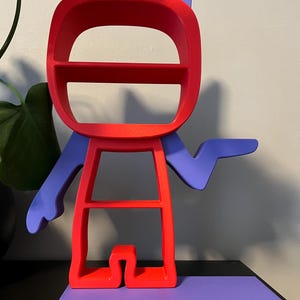 May include: A red and purple shelf unit shaped like a cartoon character. The character has a large head with two shelves, a body with one shelf, and a single arm outstretched. The character is standing on a purple base.