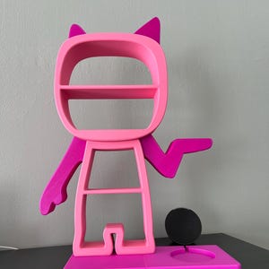 May include: A pink and magenta 3D printed shelf unit shaped like a cartoon character with cat ears. The shelf unit has three shelves and a circular cutout for a phone or other device. The base of the shelf unit is a separate piece that has a circular cutout for a phone charger.