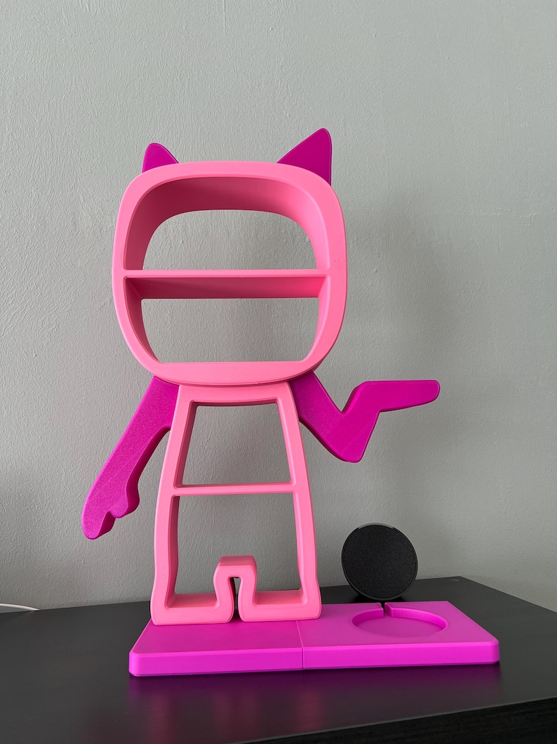 May include: A pink and magenta 3D printed shelf unit shaped like a cartoon character with cat ears. The shelf unit has three shelves and a circular cutout for a phone or other device. The base of the shelf unit is a separate piece that has a circular cutout for a phone charger.