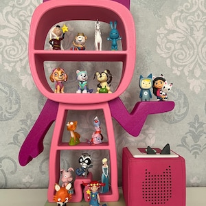 May include: A pink cat-shaped bookshelf with multiple shelves filled with small figurines. The bookshelf is standing on a colorful base with a pink speaker next to it.