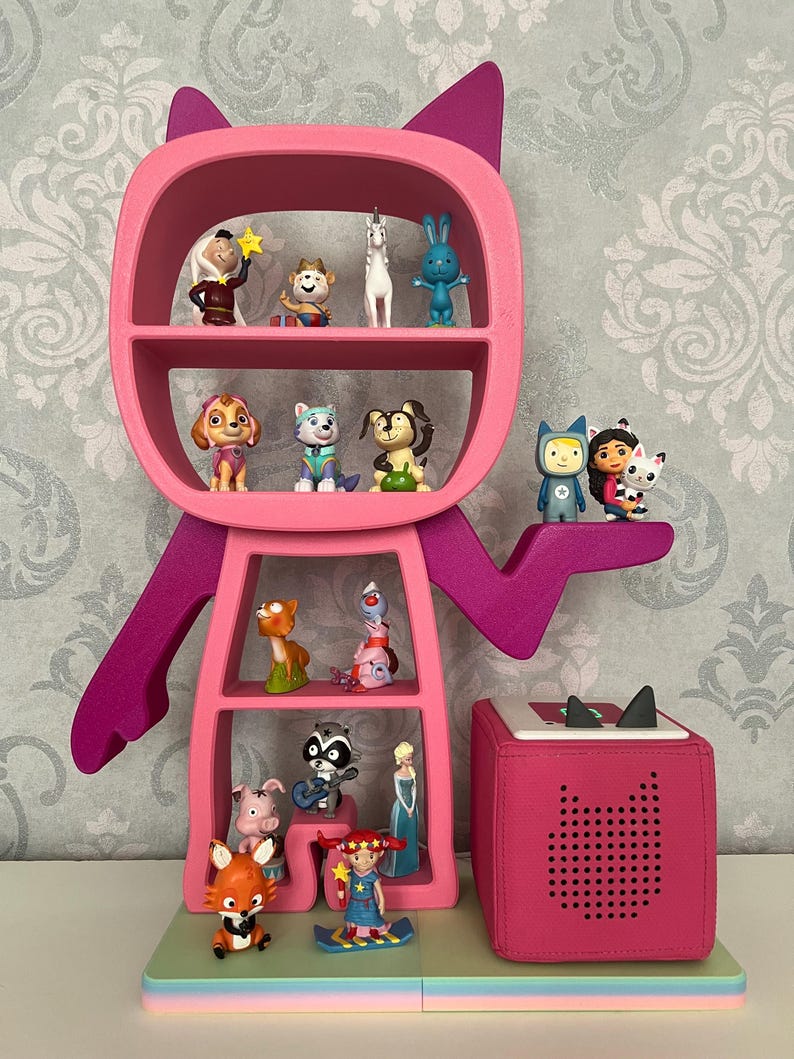 May include: A pink cat-shaped bookshelf with multiple shelves filled with small figurines. The bookshelf is standing on a colorful base with a pink speaker next to it.