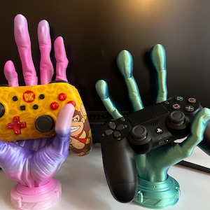 Könnte beinhalten: Zwei 3D-gedruckte Hände halten Videospielcontroller. Die linke Hand ist pink und lila und hält einen gelben und schwarzen Nintendo Switch-Controller mit dem Text "DK" darauf. Die rechte Hand ist türkis und hält einen schwarzen Playstation 4-Controller.