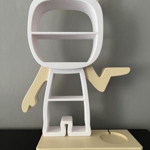 May include: White and beige shelf unit shaped like a cartoon character with cat ears. The shelf has multiple shelves and a small circular platform at the base.