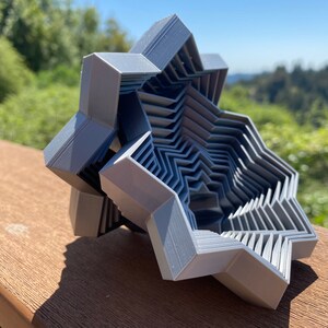 Puede incluir: Un objeto de forma de estrella impreso en 3D, de color gris, con un diseño geométrico en capas. El objeto está hecho de múltiples capas de plástico, creando un patrón complejo e intrincado.