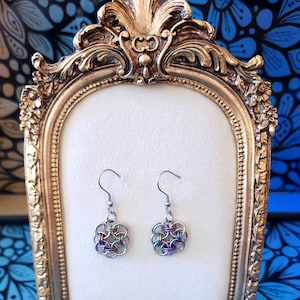 Puede incluir: Un par de pendientes de cota de malla plateados con un diseño floral y acentos iridiscentes coloridos. Los pendientes se exhiben en un marco dorado ornamentado sobre un fondo floral azul.
